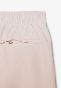 Pantoloni sportivi rosa chiaro con una vita elastica arricciata e una tasca con cerniera discreta sul retro per riporli. Texture liscia.