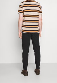 Maglietta a righe nei toni beige, nero e arancione, con scollo rotondo. Abbinata a pantaloni neri e scarpe marroni, vista da dietro.