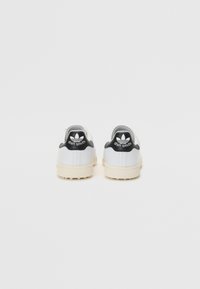 Sapatilhas brancas com parte superior em couro texturizado, pormenores pretos no calcanhar e sola em borracha creme, apresentando o logótipo "Stan Smith".