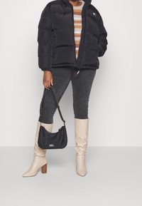 Jacke a vento nera con zip frontale, abbinata a un maglione a righe, jeans grigi, stivali beige fino al ginocchio e una piccola borsa a spalla nera.