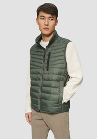 Grüne Steppweste mit Reißverschluss, horizontalen Nähten, hohem Kragen und schwarzen Akzenten. Über einem cremefarbenen Pullover getragen und mit khakifarbenen Hosen kombiniert.