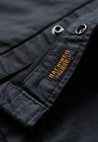 Zwart textiel met een gestructureerd oppervlak, voorzien van een oranje label met de tekst "MACHINED GOODS" en een metalen drukknopdetail.