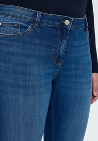 Tmavě modré džínové kalhoty s hladkou texturou, vybavené zapínáním na knoflíky, poutky na opasek a dvěma předními kapsami. Jemné šití jako detail.