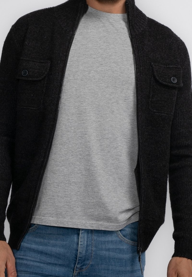 Mörkgrå stickad zip-up cardigan med två knäppta bröstfickor, i kombination med en grå T-shirt. Materialet verkar texturerat och varmt.