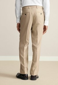 Beige Anzughose mit geradem Schnitt, ausgestattet mit zwei Gesäßtaschen mit schwarzen Knöpfen und dezenten Seitennähten. Getragen mit glänzenden schwarzen Schuhen.