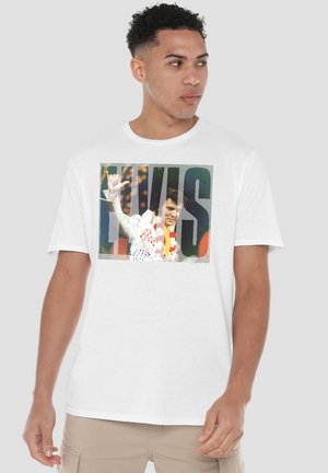 Mann trägt weißes T-Shirt mit großer bunter Elvis Presley Grafik und beigen Hosen, schaut nach links.