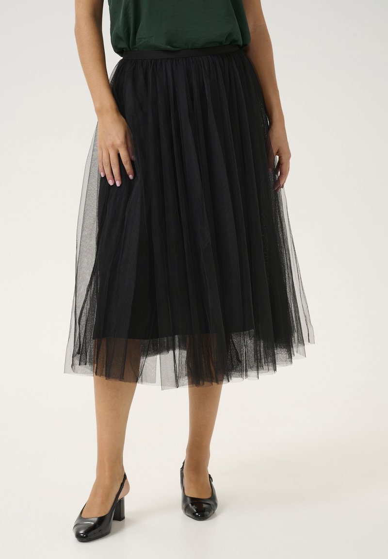 Black tulle skirt with a layered design, featuring a flexible waistband, paired with black high-heeled shoes, and a green top.
