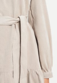 Svetlo bež obleka z vertikalnim rebrastim vzorcem, ki ima kravato okoli pasu ter naborke na rokavih in razširjen rob. Vidna je mehka tekstura.