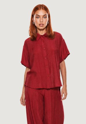Femme aux cheveux roux mi-longs, portant des boucles d’oreilles créoles argentées et une chemise rouge ample à manches courtes boutonnée avec un pantalon assorti.