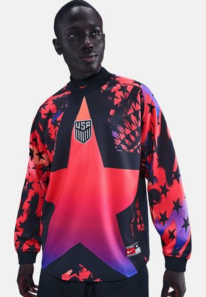 Osoba o ciemnej karnacji ubrana w czerwoną i fioletową koszulkę piłkarską USA z czarnymi gwiazdami i dużym wzorem gwiazdy z przodu.