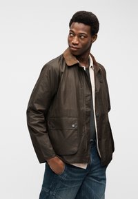 Νεαρός άνδρας με καφέ μπουφάν με γιακά από κοτλέ, μπέζ πουκάμισο και μπλε τζιν, κοιτάζει πλάγια με τα χέρια στις τσέπες, σε απλό φόντο.