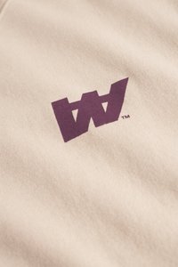 Tumši purpursarkans stilizēts "W" logo uz gaiši brūnas auduma ar smalku tekstūru un mīkstiem krokām.