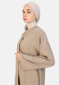 Un capo d'abbigliamento all'aperto beige chiaro a maniche lunghe con una finitura testurizzata e apertura frontale. Accompagnato da un hijab beige chiaro avvolto attorno al collo.