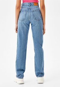 Kvinna som bär högmidjade ljusblå raka jeans med bakfickor, rosa topp och vita sneakers, stående mot vit bakgrund.