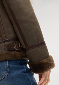 Bruine shearlingjas met een zachte bontkraag, lederen accenten en een gespdetail aan de mouw, aangevuld met een gestructureerd oppervlak.