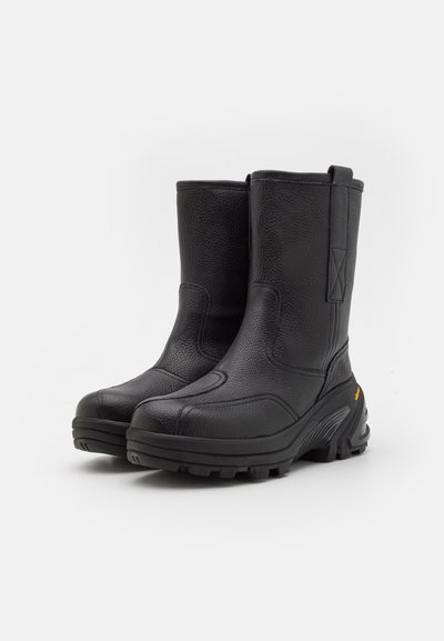 Botas de tobillo de cuero negro con acabado texturizado, puntera reforzada y suela de goma gruesa con patrones de tracción. Sin acentos de hardware visibles.