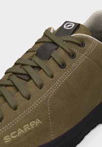 Κοντινή λήψη του sneaker σε απόχρωση ελιάς από σουέντ με κορδόνια στο ίδιο χρώμα, λευκή ραφή και μαύρη σόλα, με το λογότυπο "Scarpa" στο πλάι.