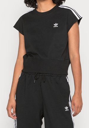Personne portant un T-shirt court noir Adidas et un pantalon assorti avec des bandes blanches et des logos sur l'épaule et la cuisse.