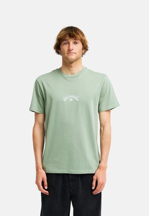 Giovane uomo con capelli castano chiaro indossa una t-shirt Billabong verde chiaro e pantaloni neri, in piedi rivolto verso l'obiettivo su uno sfondo bianco uniforme.