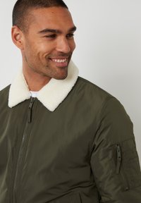 Olivgrüne Bomberjacke mit einem weißen, flauschigen Kragen, Reißverschluss und einer Seitentasche. Glatte Textur, mit gerippten Bündchen und Saum.