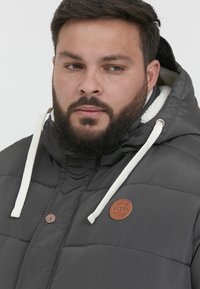 Dark gray puffer jacket with a fur-lined hood, white drawstrings, brown leather accent patch, and snap-button closure, featuring quilted texture.
