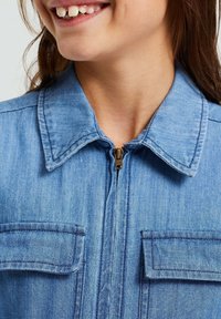 Giacca di denim di colore azzurro chiaro, con chiusura a zip, colletto a punta e due tasche sul petto con pattina. Texture morbida e design casual.