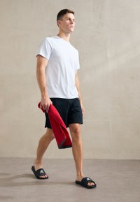 T-shirt en coton blanc, shorts noirs, sandales noires avec logo, tenant une serviette rouge, se tenant contre un mur texturé et neutre.