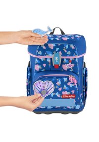 Blue backpack with heart-pattern, fabric material, pink accents, and a red logo. Hand holds a shell and a whale patch.