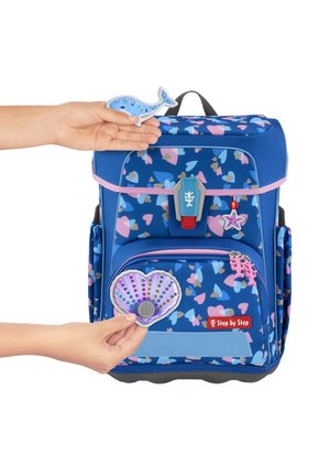 Blue backpack with heart-pattern, fabric material, pink accents, and a red logo. Hand holds a shell and a whale patch.