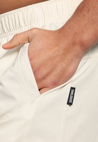 Pantalones cortos de color beige claro con cinturilla elástica y un bolsillo lateral. Presenta una etiqueta negra con "SMILODOX" en el borde del bolsillo. Textura suave.