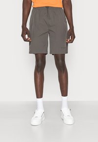 Shorts gris foncé pour hommes avec une taille élastique, un cordon de serrage, des poches latérales et des coutures plates. Portés avec des baskets blanches et des chaussettes.