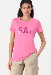 Rožinė medvilninė marškinėlių su blizgiu, ryškiu "GAP" logotipu tamsiai rožinės spalvos. Trumpomis rankovėmis, apvaliu iškirpimu ir laisvu kirpimu.