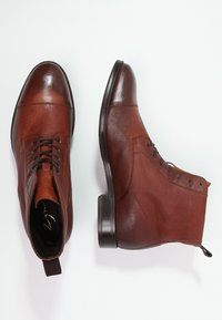 Stivali ankle in pelle marrone con finitura liscia, punta rotonda, lacci frontali e dettagli laterali. Suola nera robusta con un leggero tacco.