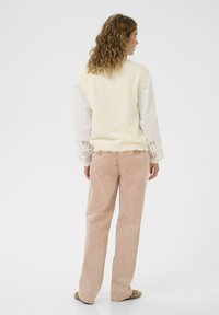 Valkoinen neulottu liivi, jossa on aaltoileva helma ja hienovaraiset kukkakuviot, päällä valkoinen paitapusero, jossa on pitsiyksityiskohtia. Vaaleanpunaiset corduropyjaman housut.