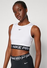 Bijela Nike Pro kratka majica s crnim pojasom na kojem piše "NIKE PRO". Glatka tkanina, uski kroj, bez rukava. Prikazane crne tajice.