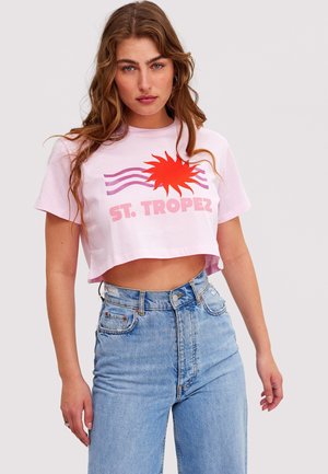 Vrouw met lang golvend haar draagt een roze cropped T-shirt met een rode zon en de tekst "ST. TROPEZ", gecombineerd met een high-waisted blauwe spijkerbroek.