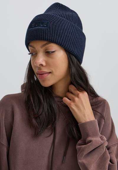 Tumši zils ribu adījuma cepure ar teksturētu rakstu un zīmolu uzlīmi. To valkā persona brūnā kapucī, atklājot matus un ādu.