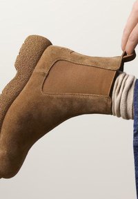 Botte chelsea en daim marron avec des panneaux élastiques sur les côtés, semelle en caoutchouc texturée et une languette; portées avec un pull gris et un jean bleu.