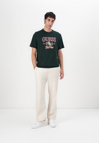 Camiseta de algodón verde oscuro con gráfico rojo y blanco de "GUESS Golf Club", combinada con pantalones de color crema y zapatillas blancas.