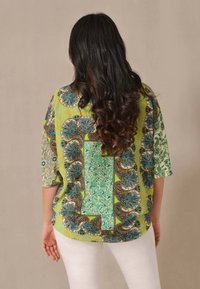 Blusa ampia gialla, verde e blu con motivi. Presenta maniche corte, una forma rilassata e disegni floreali intricati su un tessuto testurizzato.