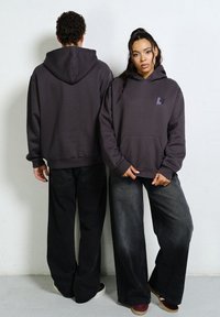 Ciemnoszare bluzy z kapturem z przednimi kieszeniami i małym logo, noszone z luźnymi czarnymi spodniami, prezentowane z przodu i z tyłu. Miękki materiał, luźny krój.