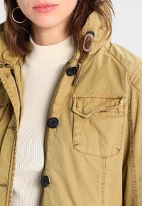 Kaki utilityjacka i bomull med dragkedja och knäppning, bröstficka och texturerade detaljer på kragen och ärmsluten.