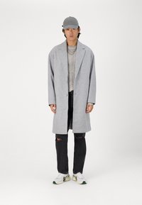 Cappotto di lana grigio con un solo bottone, indossato sopra un maglione a coste beige e jeans neri strappati. Sneakers bianche con accenti verdi.