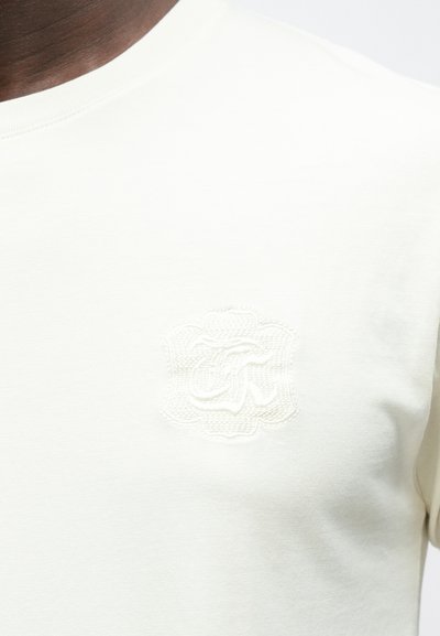 Camiseta beige claro hecha de algodón suave; presenta un logo bordado texturizado en un tono sutil. Cuello redondo estándar con mangas cortas.