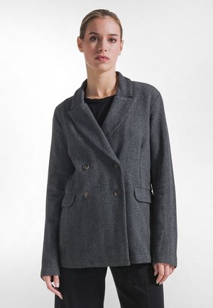 Giovane donna che indossa un blazer doppiopetto grigio con motivo a spina di pesce sopra una camicia nera, rivolta in avanti con espressione neutra.