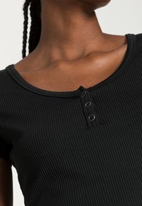 Zwarte geribbelde top met een ronde hals, drie functionele drukknoopjes en korte mouwen, met een zachte, textuur rijke finish.