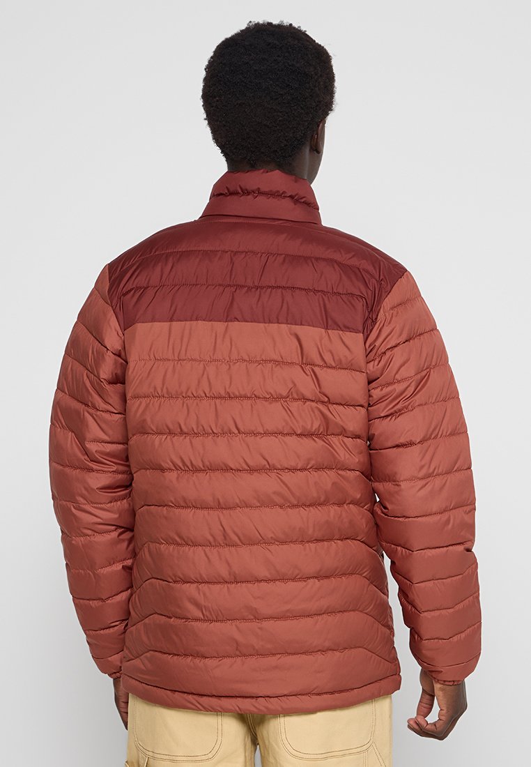 Chaqueta acolchada en tonos rojizos con un diseño acolchado horizontal. Cuenta con un cuello alto y un acabado mate, proporcionando aislamiento y calidez.