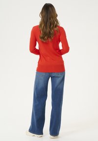 Frau mit langen, gewellten Haaren, die ein rotes langärmliges Oberteil, eine blaue weit geschnittene Jeans und weiße Turnschuhe trägt, steht vor einem schlichten Hintergrund.