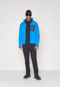 Giacca in pile blu con una tasca scura, t-shirt nera, pantaloni neri e sneakers nere. Il modello indossa un cappellino e occhiali da sole. Sfondo semplice.