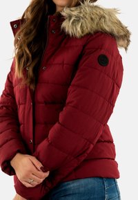 Rode donsjack met een capuchon van nepbont, met een doorgestikt ontwerp, drukknoopsluiting en een stoffen patch op de linkerarm. Gladde textuur.